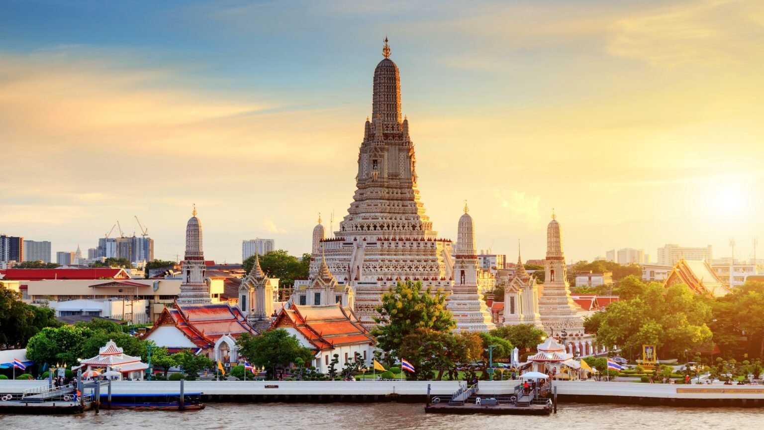 Wat Arun temple at sunset, showcasing intricate architecture and vibrant colors along the Chao Phraya River in Bangkok, Thailand. The scene includes lush greenery and traditional Thai structures, highlighting the cultural significance of this iconic landmark.