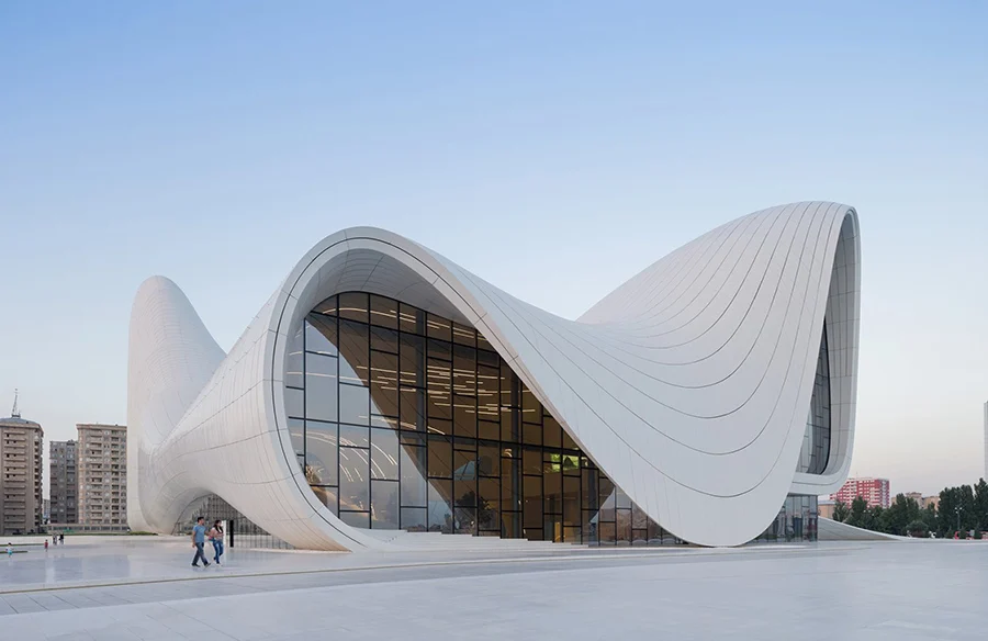 Modern architectural marvel, the Heydar Aliyev Center in Baku, Azerbaijan, features a sleek, flowing design with white curves and large glass windows, surrounded by urban landscape and clear skies.
