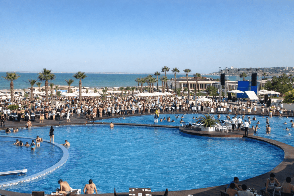 Crowded beach club scene featuring a large swimming pool, palm trees, and a vibrant atmosphere. Guests are enjoying the sun and water, with a backdrop of the ocean and event stage. Perfect for a summer getaway or social gathering.