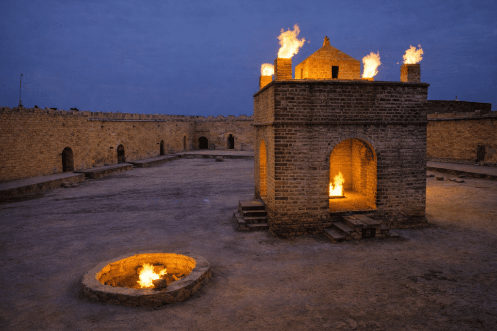 Historic fortress at dusk featuring a central structure with flames on the roof and a fire pit in the foreground, surrounded by ancient stone walls and a barren courtyard.