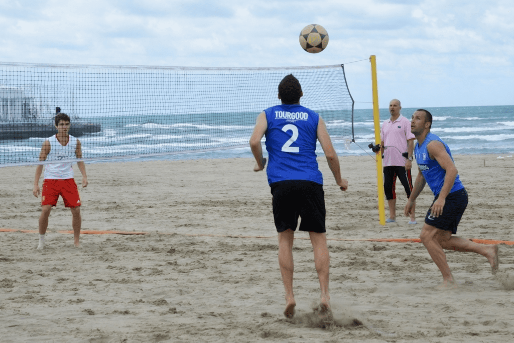 Beach soccer players engaging in a match on a sandy shoreline, with a volleyball net in the background and ocean waves visible. One player is preparing to kick the ball while others are positioned nearby, showcasing an active beach sports environment.