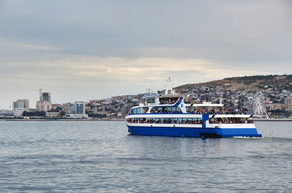 A large passenger ferry with multiple decks navigates through calm waters, with a city skyline and hills in the background, showcasing urban development and a ferris wheel.
