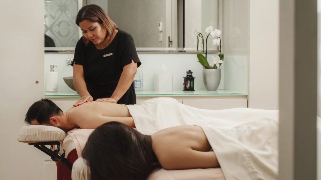 Massage therapy session in a serene spa setting, featuring a therapist providing a back massage to a male client while a female client relaxes on another massage table. The environment includes soothing decor with plants and soft lighting, promoting relaxation and wellness.