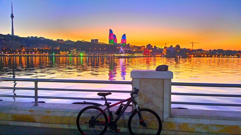 Scenic view of Baku waterfront at sunset with a bicycle parked by the railing, featuring vibrant city lights and the iconic Flame Towers in the background, reflecting in the water.