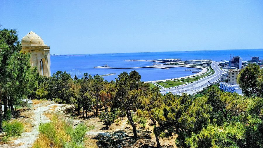 Scenic view of a coastal city featuring a historic monument, lush greenery in the foreground, and a clear blue sea in the background, showcasing modern architecture and a curved waterfront road.