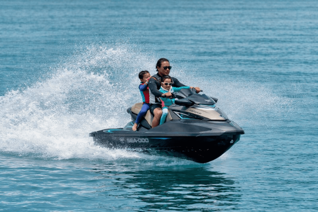 A person riding a Sea-Doo jet ski with two children, enjoying a fun day on the water, creating splashes against a serene blue ocean backdrop.