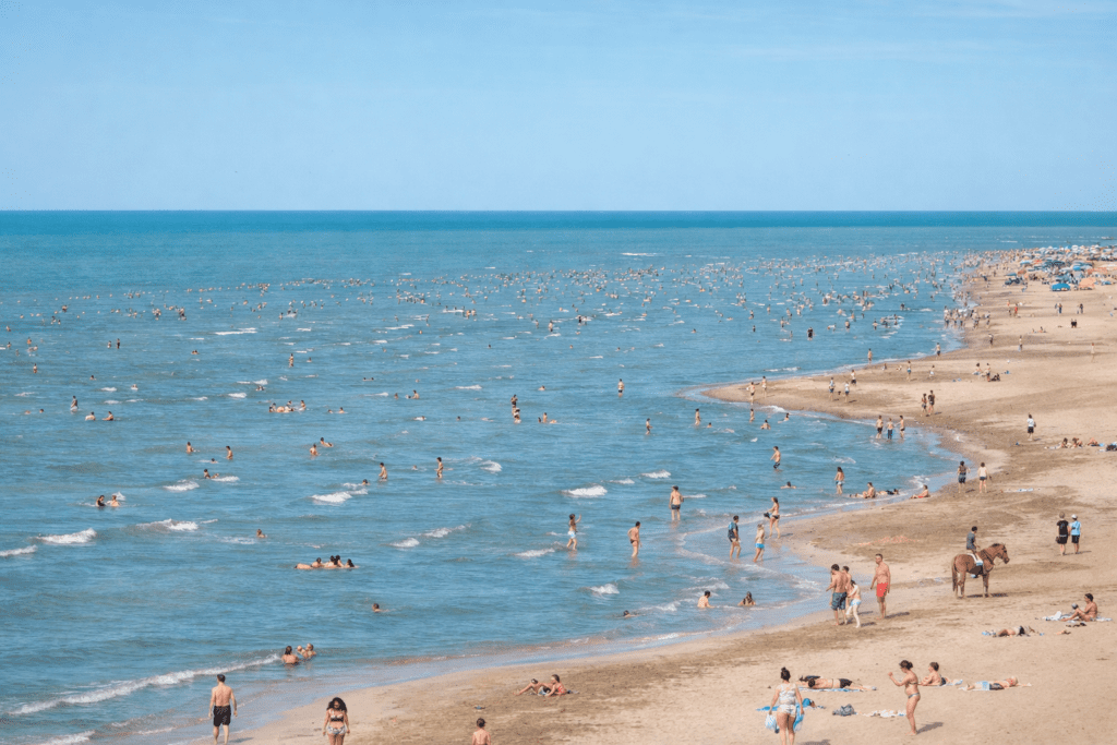 Busy beach scene with numerous people swimming and sunbathing along the sandy shore, showcasing a vibrant summer atmosphere and clear blue waters.