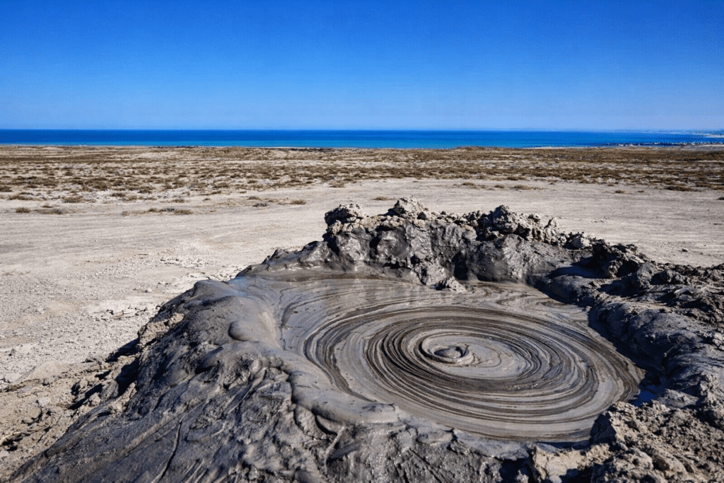 Mud volcano surrounded by arid land and a clear blue sky, with the sea visible in the background, showcasing natural geological activity.