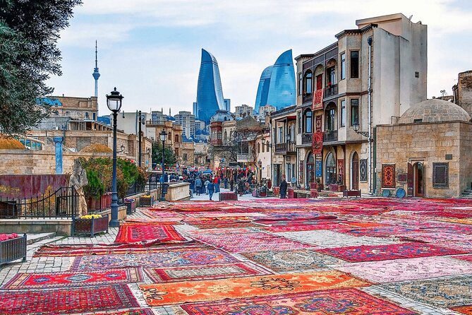 Colorful traditional rugs displayed on a cobblestone street in Baku, Azerbaijan, with modern Flame Towers in the background and local vendors and visitors exploring the area.
