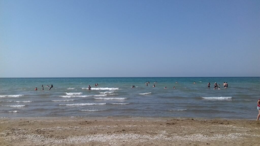 Beach scene with people swimming in the ocean under a clear blue sky, featuring gentle waves and sandy shores, ideal for summer relaxation and recreational activities.