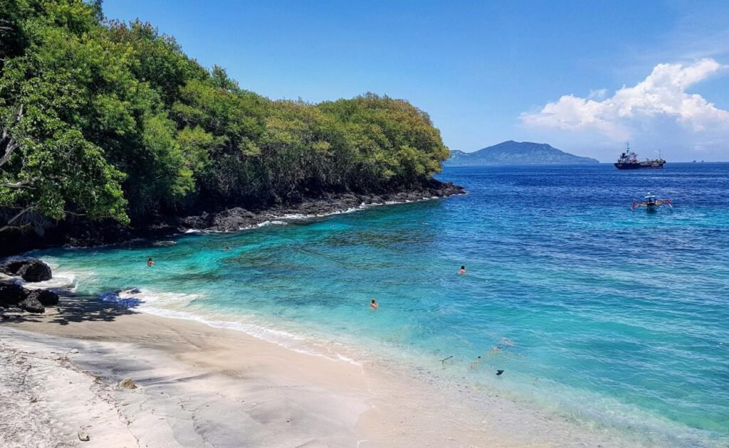 Scenic beach view featuring clear turquoise waters, sandy shore, and lush greenery along the coastline, with a boat in the distance and people swimming, ideal for tropical vacation imagery.