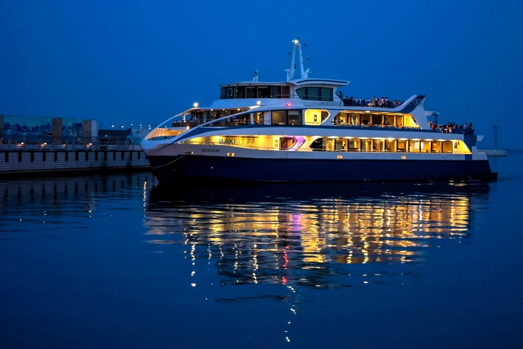 A beautifully illuminated ferry docked at a harbor during twilight, reflecting lights on the calm water, with passengers enjoying the view from the upper deck.