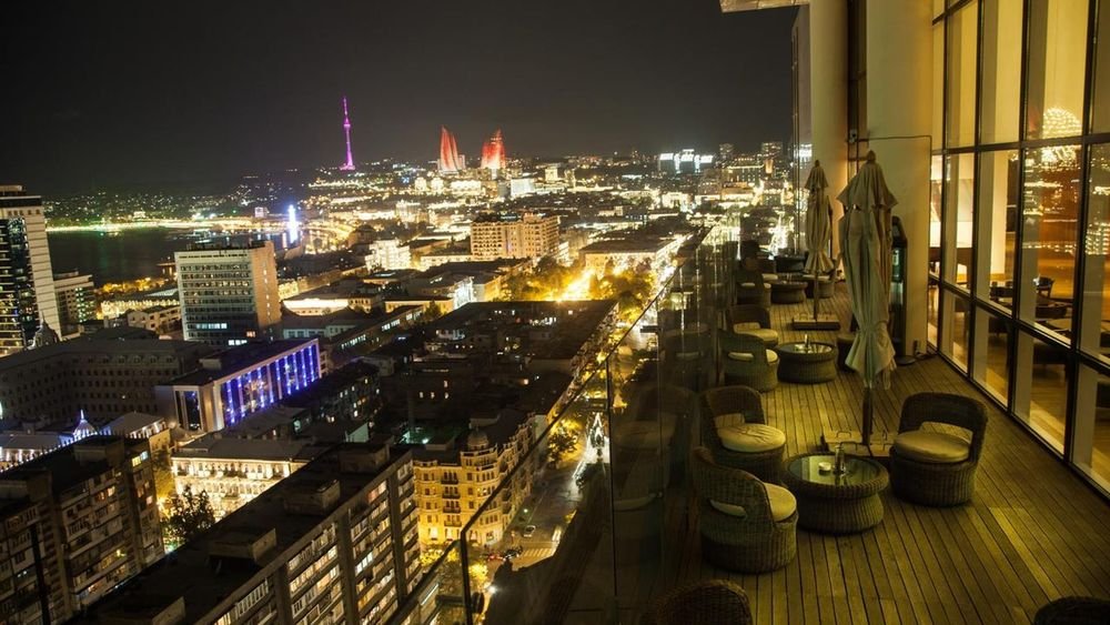 City skyline at night showcasing illuminated buildings and landmarks, including a tower with colorful lights, viewed from a modern balcony with outdoor seating.