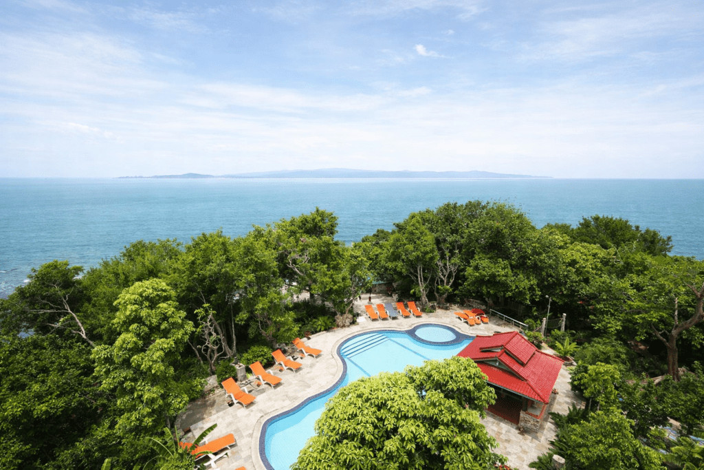Scenic view of a resort pool surrounded by lush greenery and ocean, featuring orange lounge chairs, with a backdrop of calm blue waters and distant islands under a clear sky.