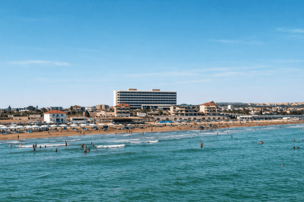 Coastal view featuring a sandy beach with sunbathers and surfers, a hotel in the background, and clear blue skies. Ideal destination for beachgoers and water sports enthusiasts.