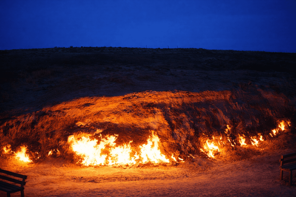 Flames erupting from a natural gas fire in a barren landscape at dusk, with a dark blue sky overhead and wooden benches in the foreground.