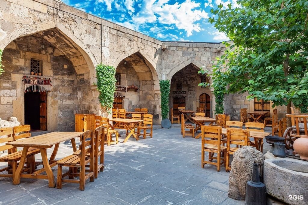 Outdoor dining area of a rustic stone courtyard featuring wooden tables and chairs, surrounded by lush greenery and historical architecture under a bright blue sky.