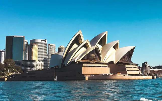 Sydney Opera House viewed from the water, showcasing its iconic sail-like architecture against a clear blue sky and the Sydney skyline in the background.