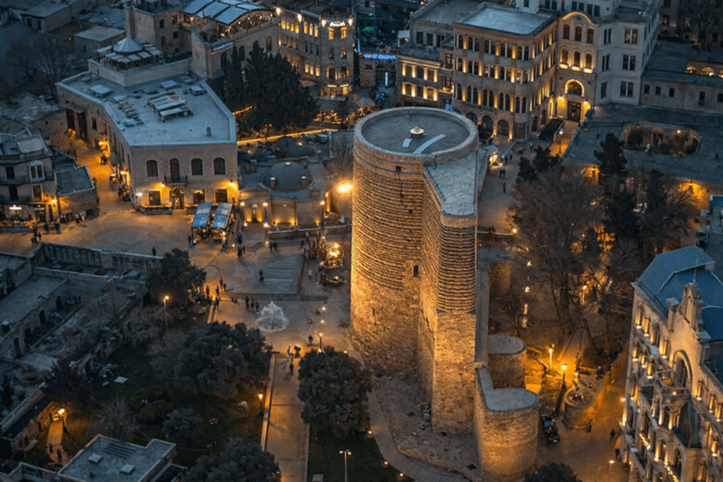 Aerial view of the Maiden Tower in Baku, Azerbaijan, surrounded by illuminated buildings and bustling streets at dusk, showcasing the blend of historical architecture and modern city life.
