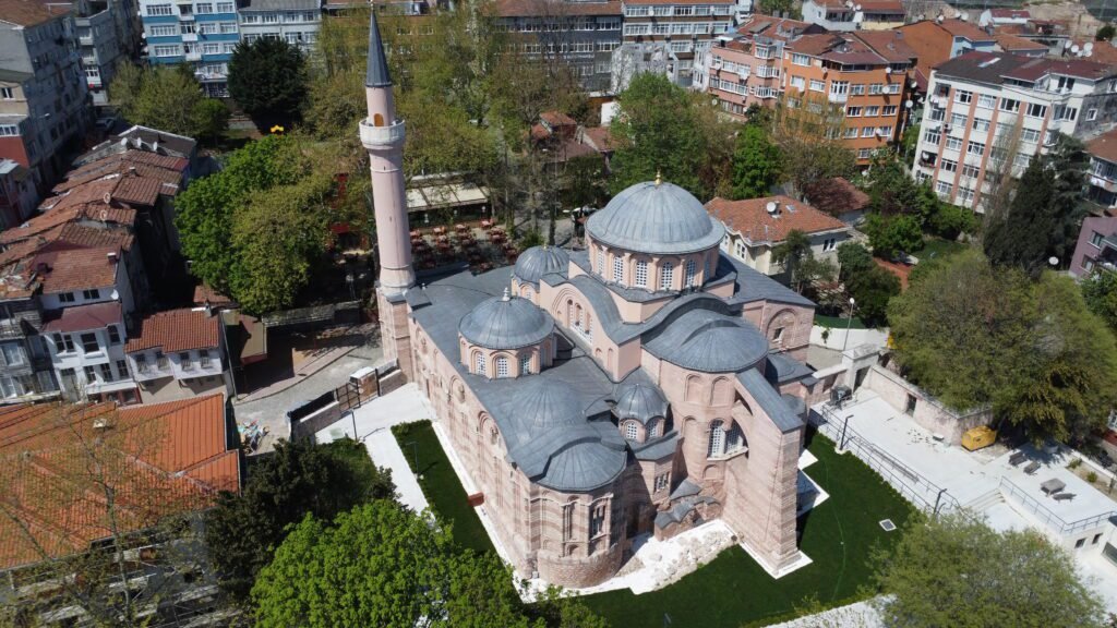 Aerial view of a historic mosque with multiple domes and a tall minaret, surrounded by greenery and residential buildings, showcasing architectural details and urban landscape.
