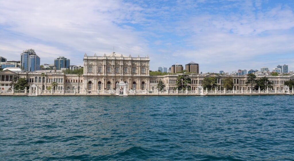 Dolmabahçe Palace in Istanbul, Turkey, showcasing its stunning architecture along the Bosphorus, surrounded by modern buildings and greenery under a partly cloudy sky.
