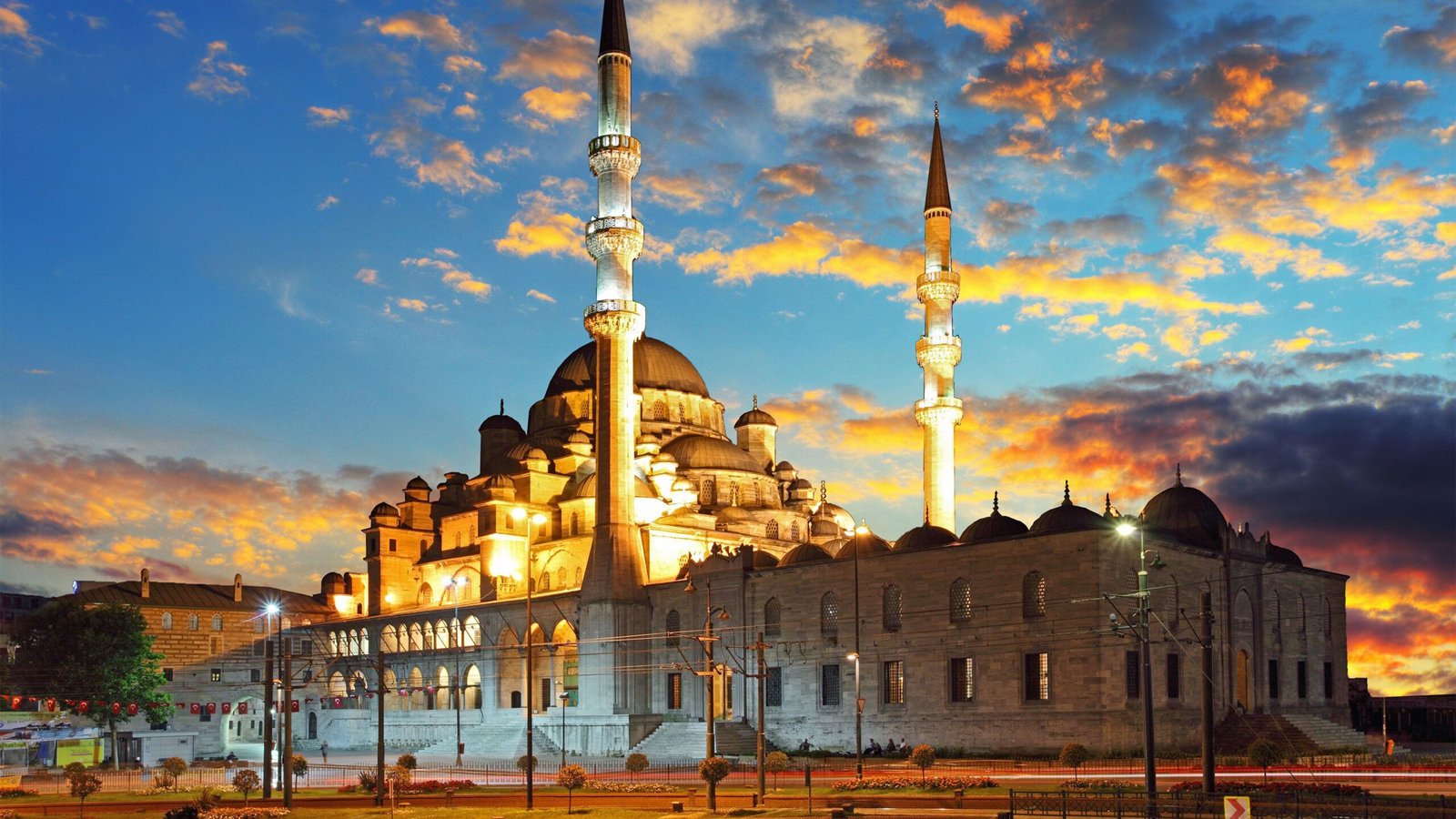 Sunset view of a grand mosque with multiple domes and tall minarets, illuminated against a colorful sky, showcasing Islamic architecture in an urban setting.
