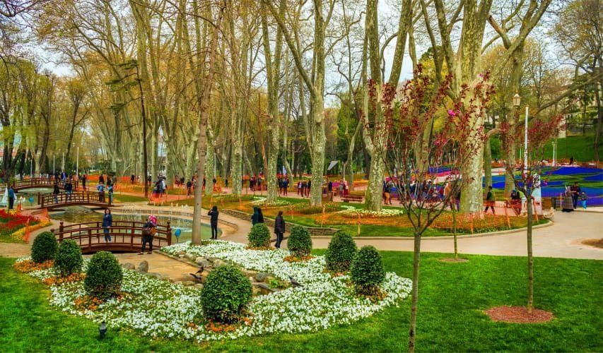A vibrant park scene featuring lush greenery, flower beds, and various visitors enjoying the outdoors. The landscape includes wooden bridges, fountains, and neatly arranged floral displays, creating a picturesque and inviting atmosphere for relaxation and leisure activities.