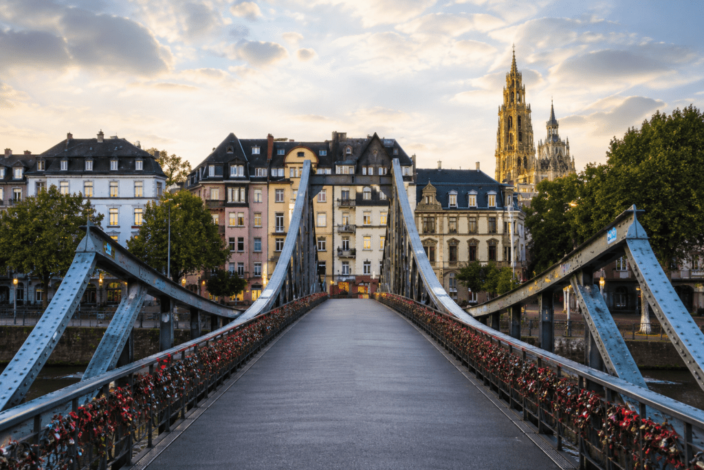 Scenic view of a bridge adorned with love locks, leading to historic buildings in Strasbourg, France, with the Strasbourg Cathedral visible in the background under a cloudy sky at dusk.