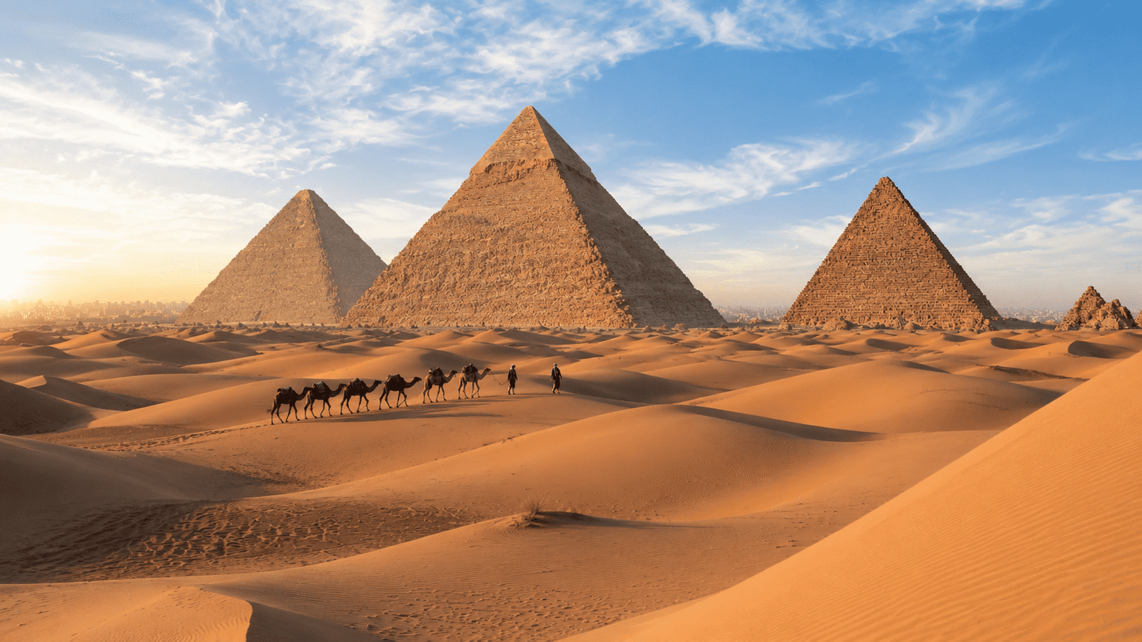 A scenic view of the Pyramids of Giza in Egypt, with a caravan of camels and travelers crossing the golden sand dunes at sunrise, showcasing the ancient architecture against a backdrop of a clear blue sky.