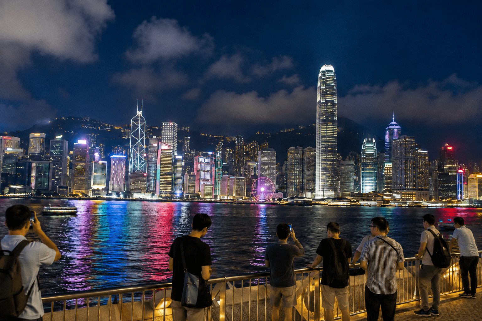 Cityscape of Hong Kong at night featuring illuminated skyscrapers and a vibrant waterfront, with several people taking photos along the promenade.