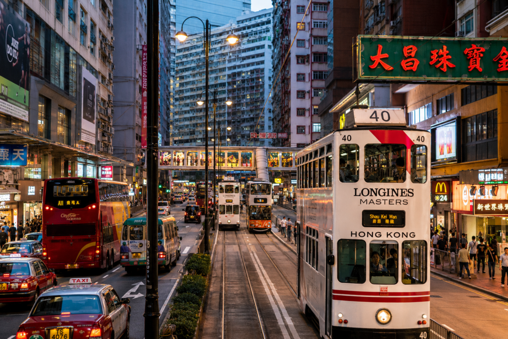 Busy street scene in Hong Kong featuring iconic double-decker trams, colorful buses, and taxis amidst towering skyscrapers and vibrant advertisements, showcasing the city's dynamic urban life.
