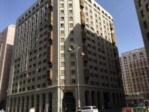 Modern hotel building in a bustling urban area, featuring a blend of architectural styles with prominent windows and decorative elements, surrounded by other structures and street activity. Ideal for travelers seeking accommodation in a central location.