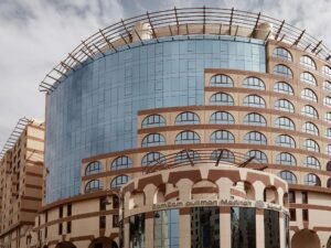 Modern architectural design of the Zam Zam Pullman Hotel in Medina, showcasing its curved glass facade and distinctive arched windows. The building features a blend of contemporary and traditional elements, with a unique rooftop structure and a welcoming entrance.