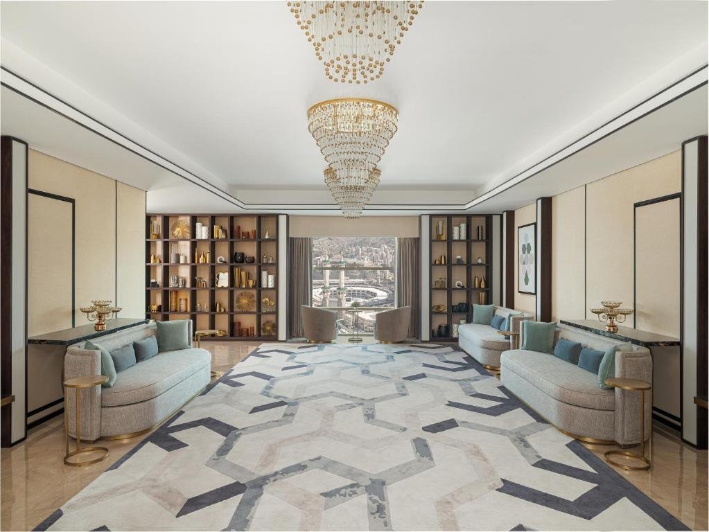 Luxurious interior of the Address Jabal Omar Makkah Hotel featuring elegant furnishings, a stunning chandelier, and a view of the city skyline through large windows. The spacious living area is designed with a modern aesthetic, showcasing a stylish rug and decorative shelving.