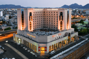 Madinah Mövenpick Hotel exterior view at dusk, showcasing its modern architecture and illuminated signage, surrounded by urban landscape and mountains in the background.