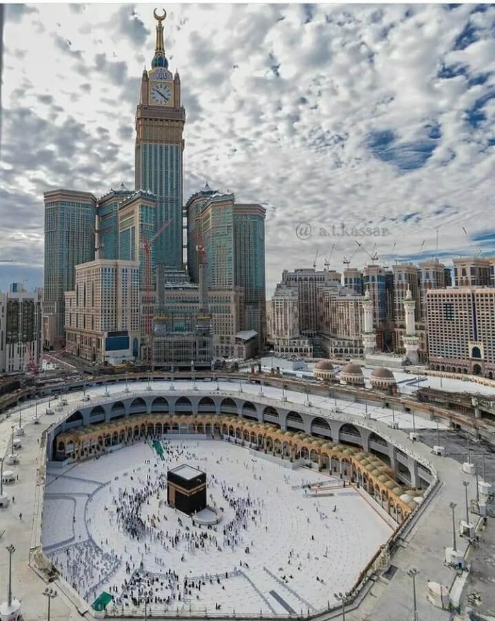 Aerial view of the Grand Mosque in Mecca, featuring the Kaaba at the center surrounded by worshippers, with the Abraj Al Bait Towers towering in the background under a cloudy sky.