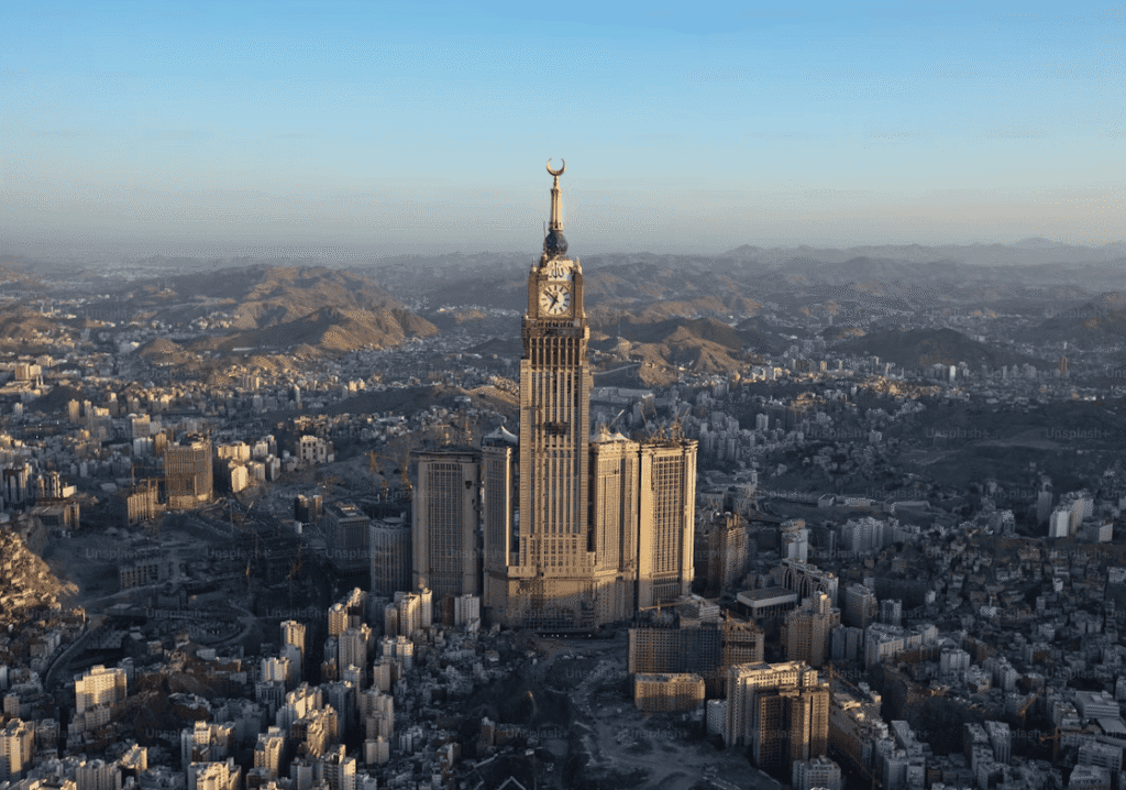 Aerial view of the Abraj Al Bait Towers in Mecca, Saudi Arabia, showcasing the iconic clock tower surrounded by urban development and mountainous terrain under a clear sky.