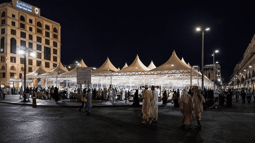 Crowd of people walking near the Dallah Taibah Hotel at night, with illuminated tents and vibrant street lighting in a bustling marketplace setting.