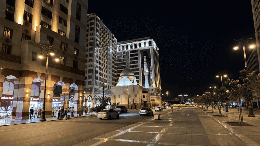 Night view of a bustling street featuring modern buildings and traditional architecture, illuminated by streetlights. The scene includes shops with visible signage, pedestrians, and vehicles on the road, showcasing urban life in a vibrant city atmosphere.