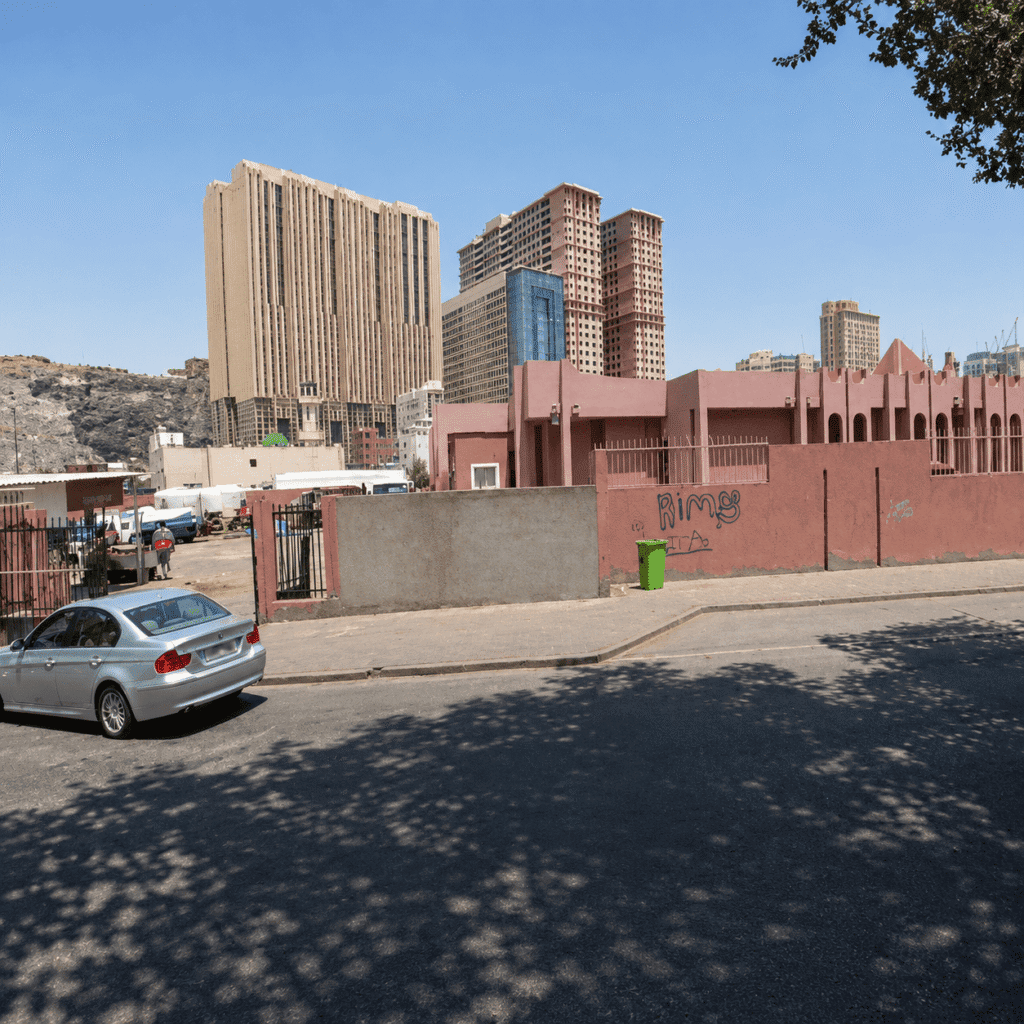 Cityscape featuring modern high-rise buildings and a pink-walled structure with graffiti, alongside a parked silver car on a sunny day.