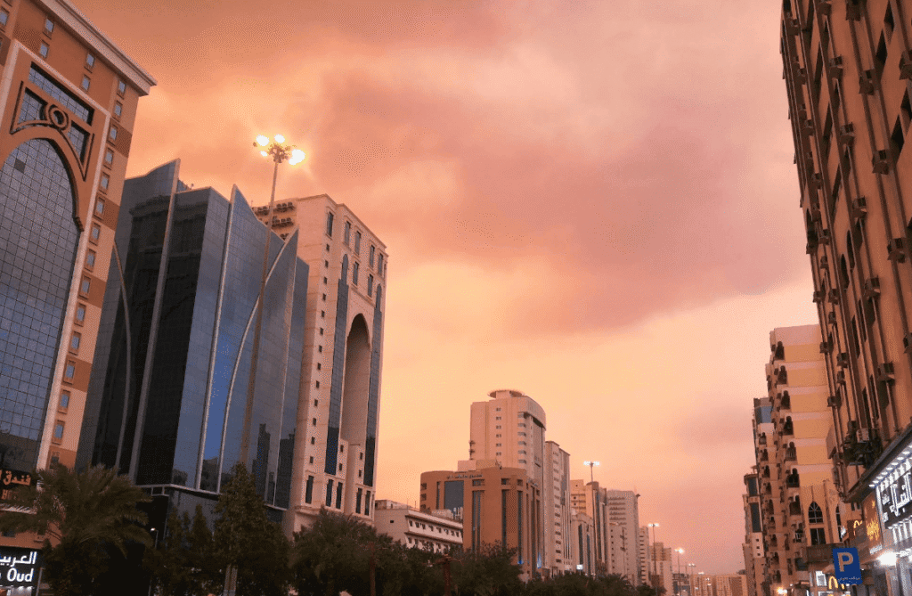 City skyline during sunset with glowing clouds, featuring modern buildings and palm trees lining the street.