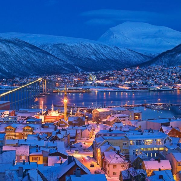 Snow-covered town illuminated at night, with mountains in the background and a bridge spanning the water, showcasing a serene winter landscape.