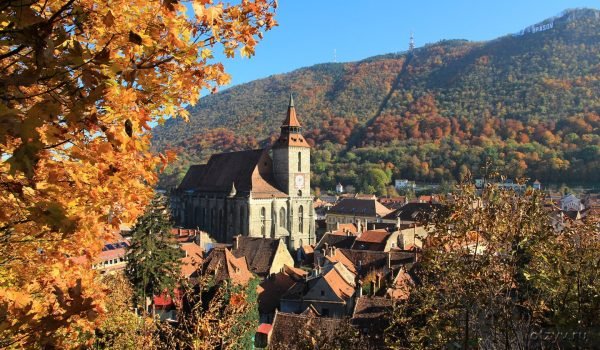 Scenic view of a historic church surrounded by autumn foliage and colorful rooftops in a picturesque town, with rolling hills in the background under a clear blue sky.