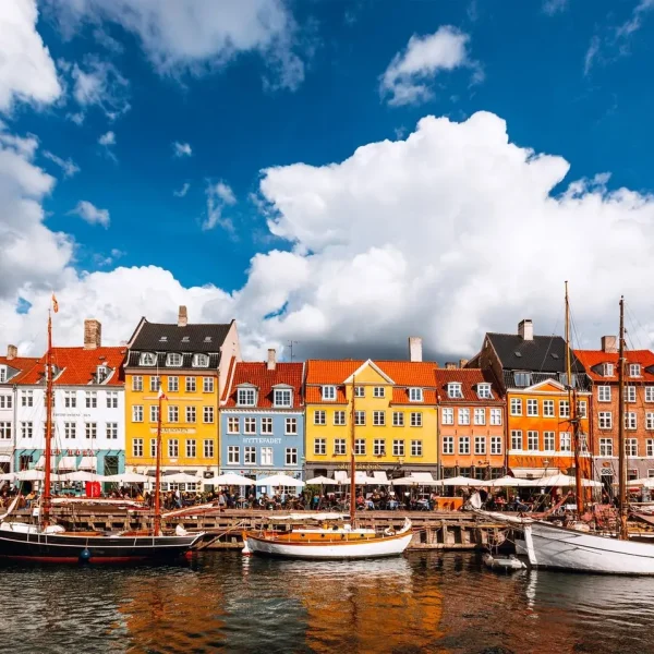 Colorful waterfront buildings in Nyhavn, Copenhagen, with boats docked along the canal under a bright blue sky and fluffy white clouds.