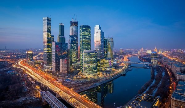 Aerial view of a modern city skyline at dusk, featuring tall skyscrapers and a river with a busy bridge illuminated by city lights.