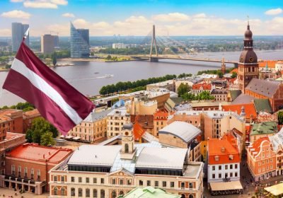 Aerial view of Riga, Latvia, showcasing the vibrant cityscape with historic architecture, the Daugava River, and the Latvian flag proudly displayed in the foreground against a clear blue sky.