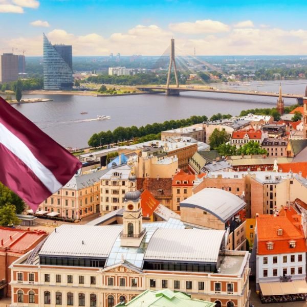 Aerial view of Riga, Latvia, showcasing the vibrant cityscape with historic architecture, the Daugava River, and the Latvian flag proudly displayed in the foreground against a clear blue sky.