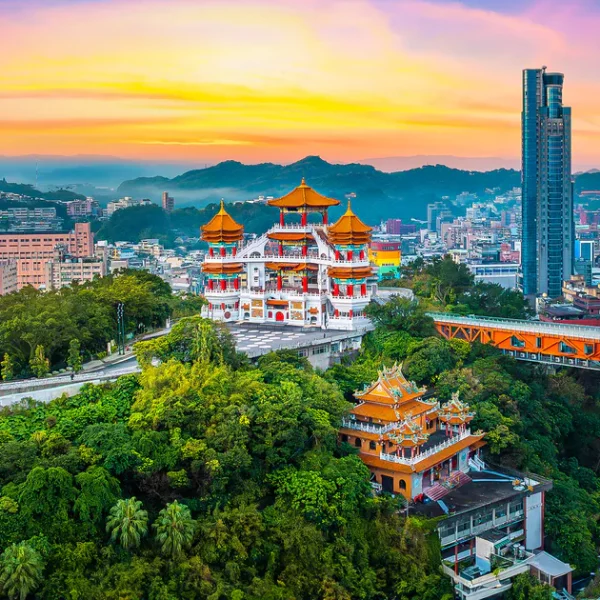 Aerial view of a traditional temple surrounded by lush greenery at sunset, with modern skyscrapers in the background, showcasing the contrast between nature and urban architecture.