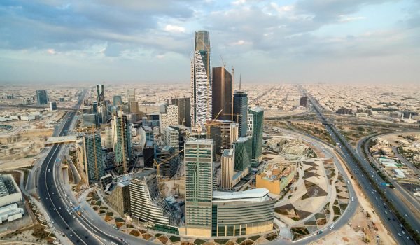 Aerial view of Riyadh's skyline featuring modern skyscrapers, including the iconic Kingdom Centre, surrounded by desert landscape and urban development.
