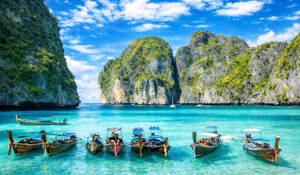 Thailand Tropical beach scene featuring traditional longtail boats anchored in crystal-clear turquoise waters, surrounded by lush green cliffs and a bright blue sky with scattered clouds. Ideal for travel and nature enthusiasts.